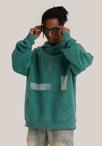 Produktfoto mit Link: Männliches dunkelhäutiges Model von vorn trägt einen grünen übergroßen Kapuzenpullover. Aufnahme bis zu den Oberschenkeln. Der Mann steht vor neutral beigem Hintergrund. Der Pullover ist dezent mit wenigen unterschiedlich langen Längs- und Querstreifen bedruckt, die Ausschnitte eines alten Posters von 1961 des US Government Printing Office zeigen. Das Poster zeigt abstrakte Wolkenkratzer-Formationen in Blau-Grün-Gelbgold. Auf der Brust prangt das gestickte Logo von Places/Pieces in Goldgelb. Produkt namens 