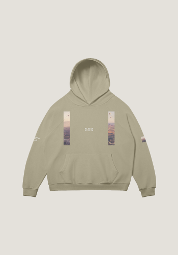 Produktfoto mit Link: Flatlay-Aufnahme eines kamelfarbenen übergroßen Kapuzenpullovers von vorn vor neutral-beigem Hintergrund. Der Pullover ist dezent mit zwei Längsstreifen links und rechts bedruckt, der Ausschnitte der unwirklichen Felslandschaft in Kappadokien in der Türkei zeigt. Über dem Boden schweben Ballons. Die Streifen changieren in Braun-Orange-Aprikose-Tönen. Auf Brusthöhe ist das Places/Pieces-Logo in weiß aufgestickt. Produkt namens 