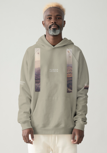 Produktfoto mit Link: Männliches dunkelhäutiges Model von vorn trägt einen kamelfarbenen übergroßen Kapuzenpullover. Aufnahme bis zu den Oberschenkeln. Der Mann steht vor neutral beigem Hintergrund. Der Pullover ist dezent mit zwei Längsstreifen links und rechts bedruckt, der Ausschnitte der unwirklichen Felslandschaft in Kappadokien in der Türkei zeigt. Über dem Boden schweben Ballons. Die Streifen changieren in Braun-Orange-Aprikose-Tönen. Auf Brusthöhe ist das Places/Pieces-Logo in weiß aufgestickt. Produkt namens 