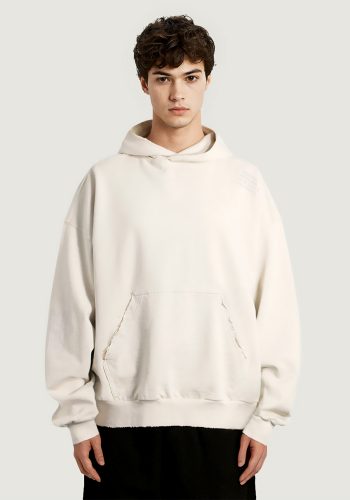 Produktfoto mit Link: Front-Aufnahme eines dunkelhaarigen Mannes, der einen ausgefransten, stylischen beigen Kapuzenpullover trägt. Auf dem Ärmel und der Schulter ist ein schlichter Text zu sehen über Amsterdam. Der Text ist Ton in Ton und nur leicht zu sehen.