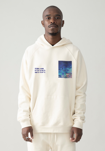 Produktfoto mit Link: Männliches dunkelhäutiges Model von vorn trägt einen hellbeigen Kapuzenpullover mit warmem Gelbstich. Aufnahme bis zu den Oberschenkeln. Der Mann steht vor neutral beigem Hintergrund. Der Kapuzenpullover zeigt die Küstenregion der Stadt 