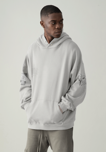 Produktfoto mit Link: Dunkelhäutiges männliches Model trägt einen hellgrauen dicken Kapuzenpullover mit Druckknöpfen am Hals. Der Mann steht vor neutral beigem Hintergrund und hat die Hände in der Kängurutasche. Der Kapuzenpullover ist auf den Ärmel mit Mondmotiven bedruckt und auf der linken Brustseite ist ein Text zu den abgebildeten Orten aufgedruckt. Produkt namens 