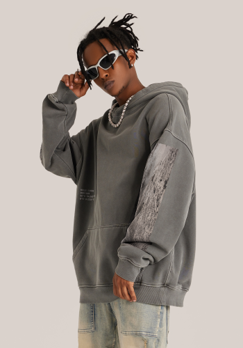 Produktfoto mit Link: Dunkelhäutiges männliches Model trägt einen mittelgrauen Kapuzenpullover. Aufnahme halb seitlich bis zu den Oberschenkeln. Der Mann steht vor neutral beigem Hintergrund und greift sich an seine weiße Sonnenbrille. Der Kapuzenpullover ist auf den Ärmel mit beige-grauen Strandmotiven aus Hvide Sande in Dänemark bedruckt (je ein langer Streifen). Links auf der Brust sind die Koordinaten des Ortes aufgedruckt. Produkt namens 