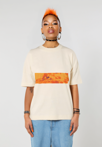 Weibliches dunkelhäutiges Model mit orange gefärbten Haaren von vorn trägt ein beiges übergroßes T-Shirt. Aufnahme bis zu den Oberschenkeln. Die Frau steht vor neutral grauem Hintergrund. Auf dem T-Shirt ist ein knalllig orangenes Motiv auf Höhe der Brust aufgedruckt. Der Querstreifen zeigt den stilisierten Ausbruch des Vulkans Mount St. Helen 1980 in den USA. Produkt namens 