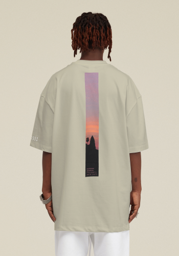 Produktfoto mit Link: Männliches dunkelhäutiges Model mit kurzen Rastas von hinten trägt ein beiges übergroßes T-Shirt. Aufnahme bis zu den Oberschenkeln. Der Mann steht vor neutral grauem Hintergrund. Auf dem T-Shirt erstreckt sich ein langer Längs-Streifen von oben nach unten in Orange, Lila und Schwarz. Er zeigt einen Ausschnitt der Tempelanlage in Prambanan in Indonesien. Produkt namens 