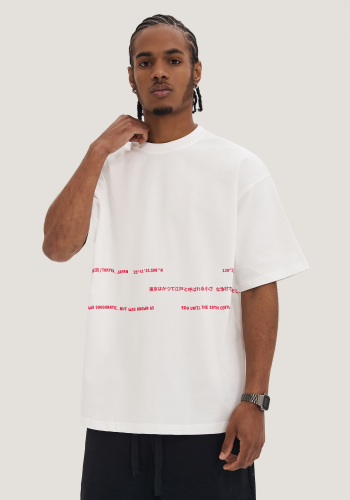 Produktfoto mit Link: Männliches dunkelhäutiges Model von vorn vor neutral-beigem HIntergrund trägt ein weißes übergroßes T-Shirt mit roten Textmotiven, teils auf japanisch, die sich quer über das T-Shirt schlängeln. Die Texte erstrecken sich auf sehr dezente Art und Weise über drei Reihen, sind versetzt zueinander angeordnet und behandeln historische Infos zu Tokio in Japan. Produkt namens 