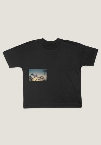 Produktfoto mit Link: Flatlay-Aufnahme eines übergroßen schwarzen T-Shirts von vorn mit hochabschließendem Kragen vor neutral-beigem Hintergrund. Auf dem T-Shirt ist auf der rechten Seite auf Brusthöhe ein rechteckiges Motiv mit Basketball-Korb in tropischer Palmenlandschaft in Los Angeles bei Abendrot abgebildet. Produkt namens 