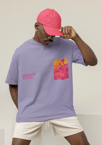 Produktfoto mit Link: Dunkelhäutiges männliches Model mit pinker Baseball-Cap sitzt lässig auf einem beigen Würfel im Fotostudio. Er trägt ein lilafarbenes Oversize-T-Shirt. Auf der linken Brust-Seite ist ein verfremdeter Kaktus in Gelb und Pink zu sehen (Viereck). Auf der anderen Seite sieht man die Koordinaten des Ortes in Pink. Produkt namens 
