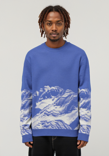 Produktfoto mit Link: Männliches dunkelhäutiges Model von vorn trägt einen sattblauen Strickpullover mit einem eingearbeiteten Motiv, das um den ganzen Pullover geht. Aufnahme bis zu den Oberschenkeln. Der Mann steht vor neutral beigem Hintergrund. Der Pullover zeigt den Gipfel des Columbus Glätschers und den Mount St. Elias in Alaska in Weiß. Das Motiv erstreckt sich von unten bis zur Mitte des Pullovers ringsrum. Produkt namens 