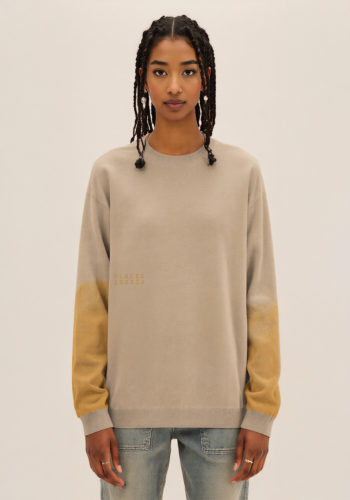 Produktfoto mit Link: Weibliches dunkelhäutiges Model von vorn trägt einen sandig-beigen Strickpullover mit einem eingearbeiteten Motiv, das um den ganzen Pullover geht. Aufnahme bis zu den Oberschenkeln. Die Frau stehet vor neutral beigem Hintergrund. Der Pullover zeigt die Baum-Landschaft am Brenner Pass zwischen Österreich und Italien in Gelb auf den beiden Ärmeln. Weil das Motiv aber in die Strick-Textur eingearbeitet ist, sieht es fast nur aus wie ein gelber Verlauf, der oben ausfranst. Vorne, auf der rechten Seite, ist noch das Places/Pieces-Logo eingearbeitet. Produkt namens 