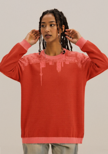 Produktfoto mit Link: Weibliches dunkelhäutiges Model von vorn trägt einen Strickpullover in sattem Rot mit korallenfarbenen Ärmeln und Kragen (korallenfarben ist ein mattes, mittleres Rosarot). In den Pullover ist Motiv eingearbeitet, das oben um den ganzen Pullover geht. Aufnahme bis zu den Oberschenkeln. Die Frau steht vor neutral beigem Hintergrund. Der Pullover zeigt die Skyline von Toronto, aber auf den Kopf gestellt, im beschriebenen Korallen-Rosarot. Das Motiv geht von vorn über beide Ärmel bis nach hinten. Produkt namens